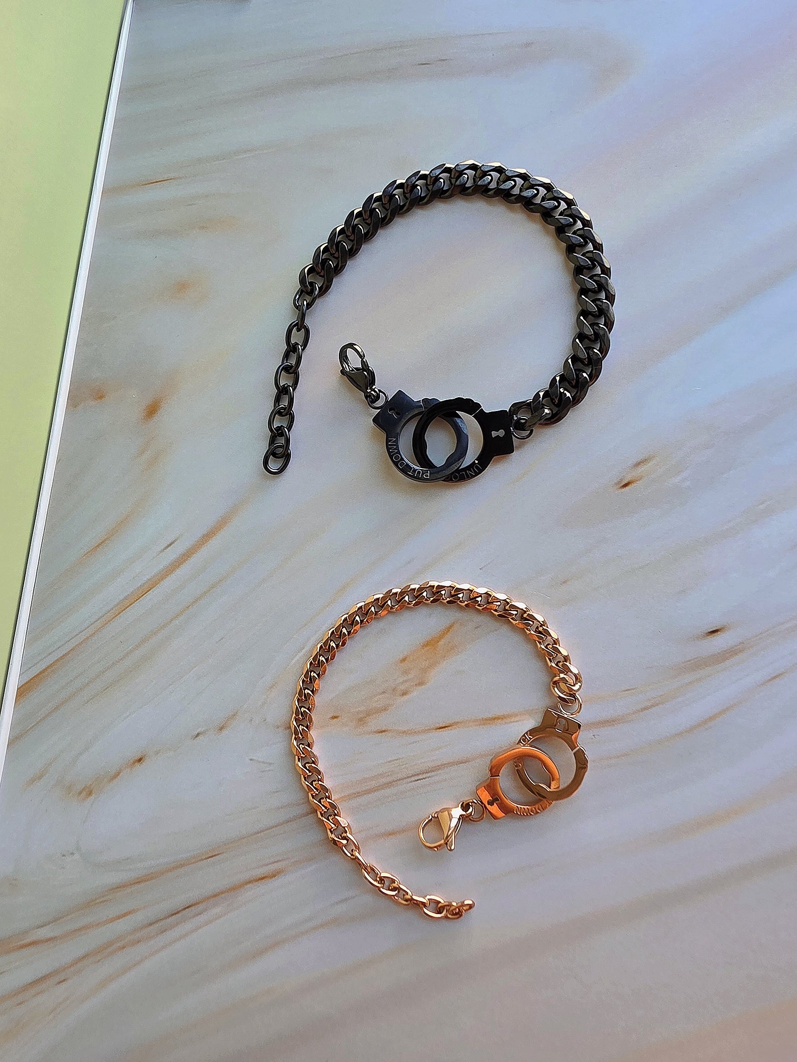 Two chain bracelets, one black and one rose gold, on a light marble surface.