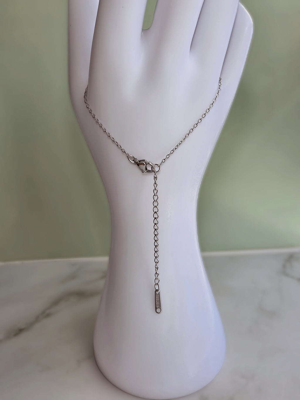 A silver-toned delicate chain necklace displayed on a white hand-shaped jewelry stand, featuring a small clasp and a dangling chain with a tiny tag at the end, set against a soft green background.