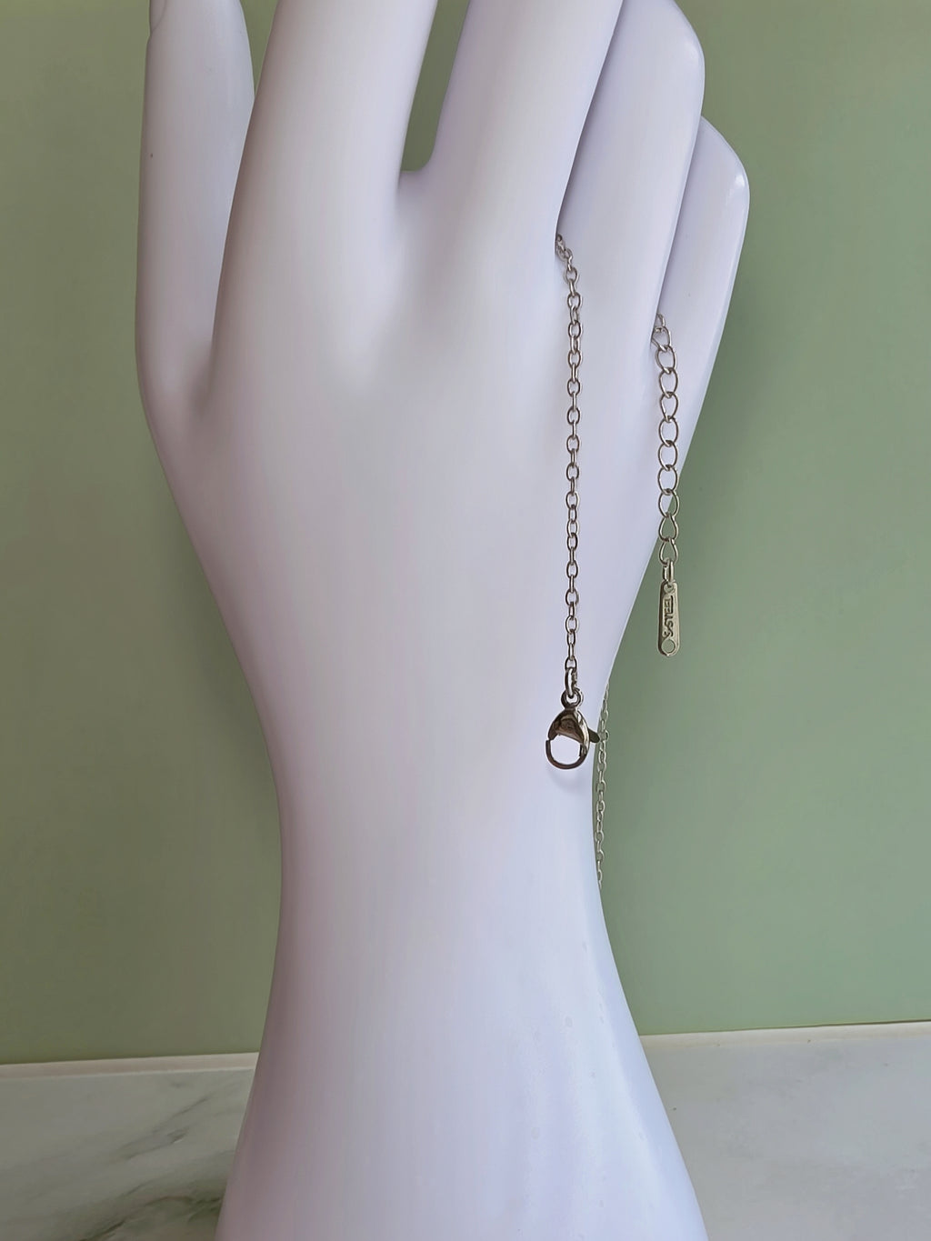 A silver-toned chain bracelet with a small ring charm and a textured dangling tag, displayed on a white hand-shaped mannequin against a pale green background.
