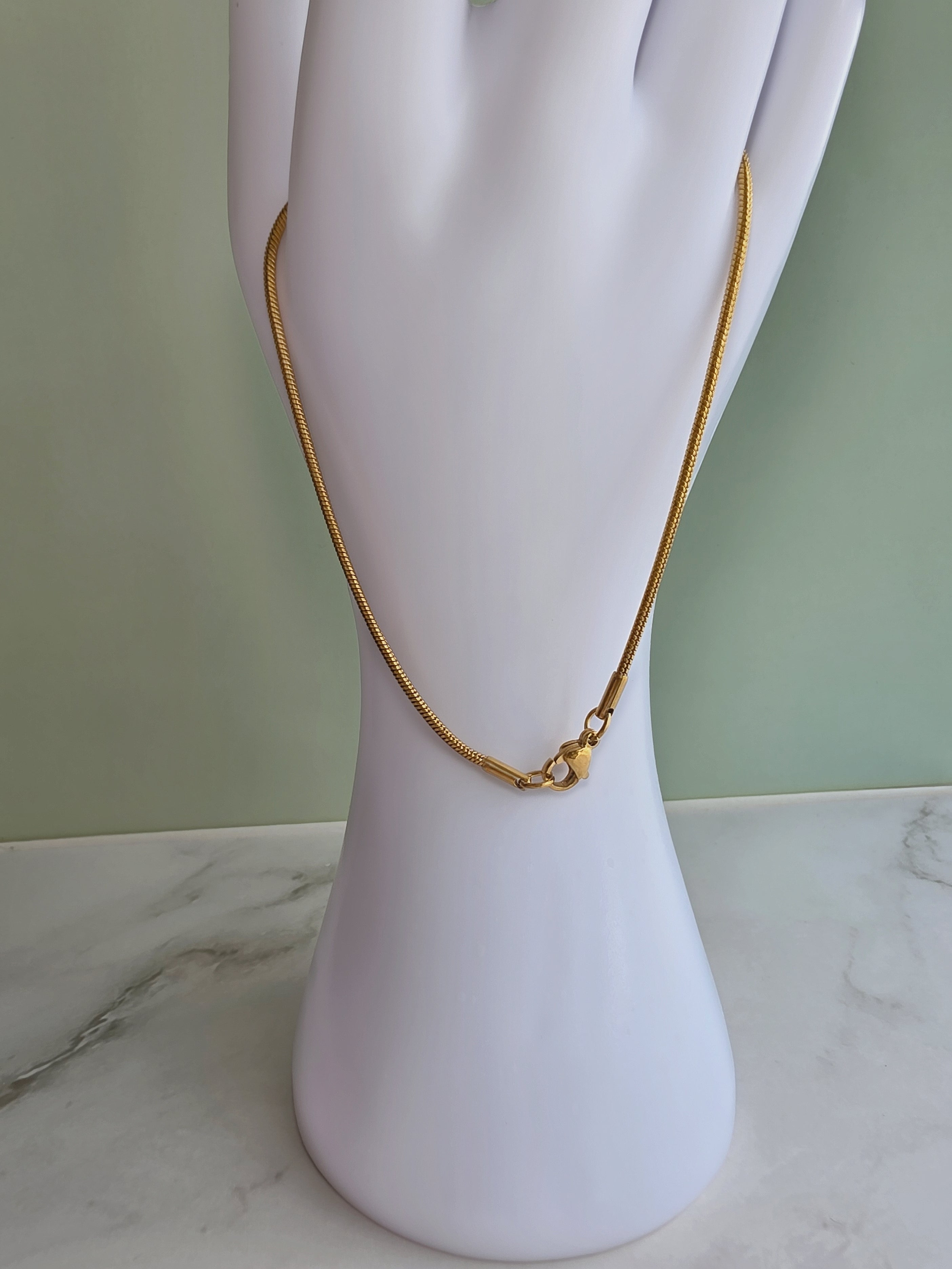 A gold chain necklace with a small gold bead pendant, displayed on a white mannequin hand against a pale green wall and marble surface.
