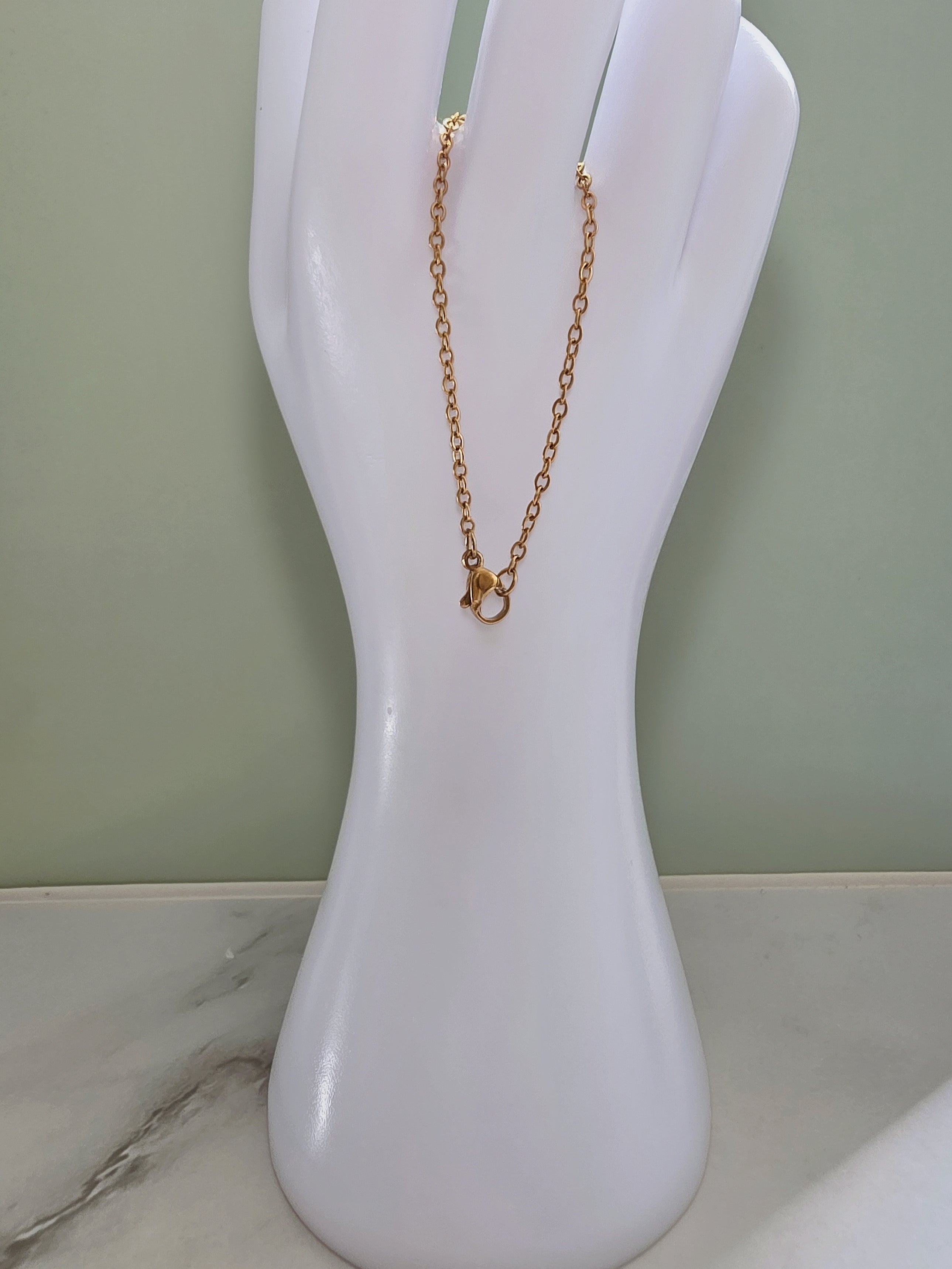 A delicate gold chain necklace with a small ring charm, displayed on a white hand-shaped jewelry stand against a light green wall.