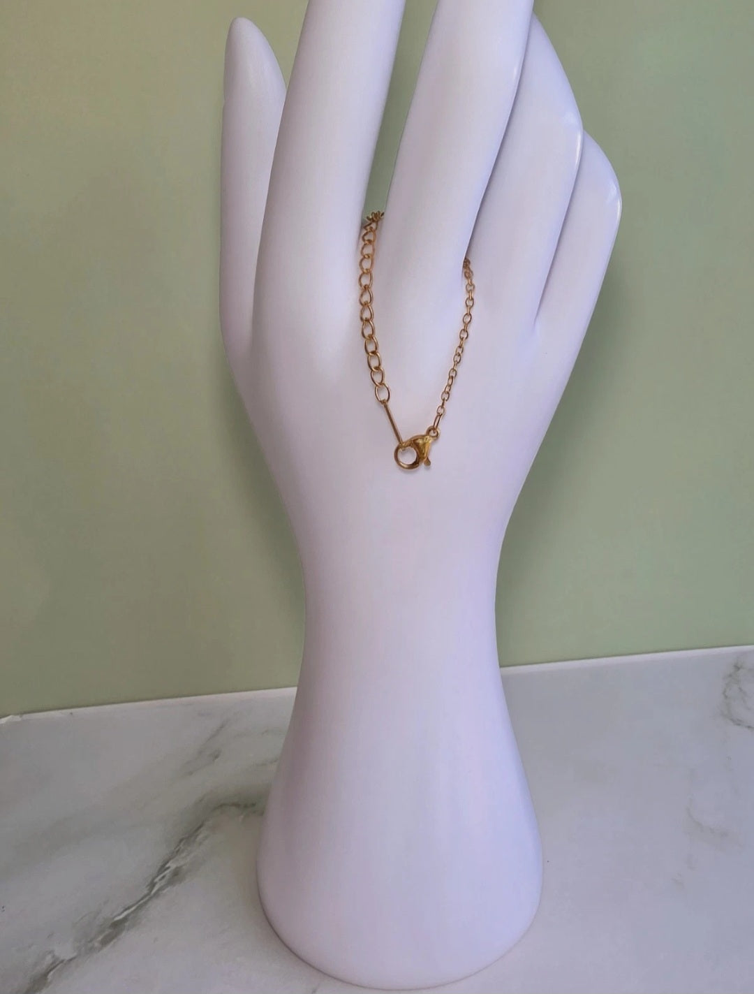 A delicate gold chain bracelet with a small lobster clasp, displayed on a white hand-shaped jewelry stand against a pale green wall and marble surface.
